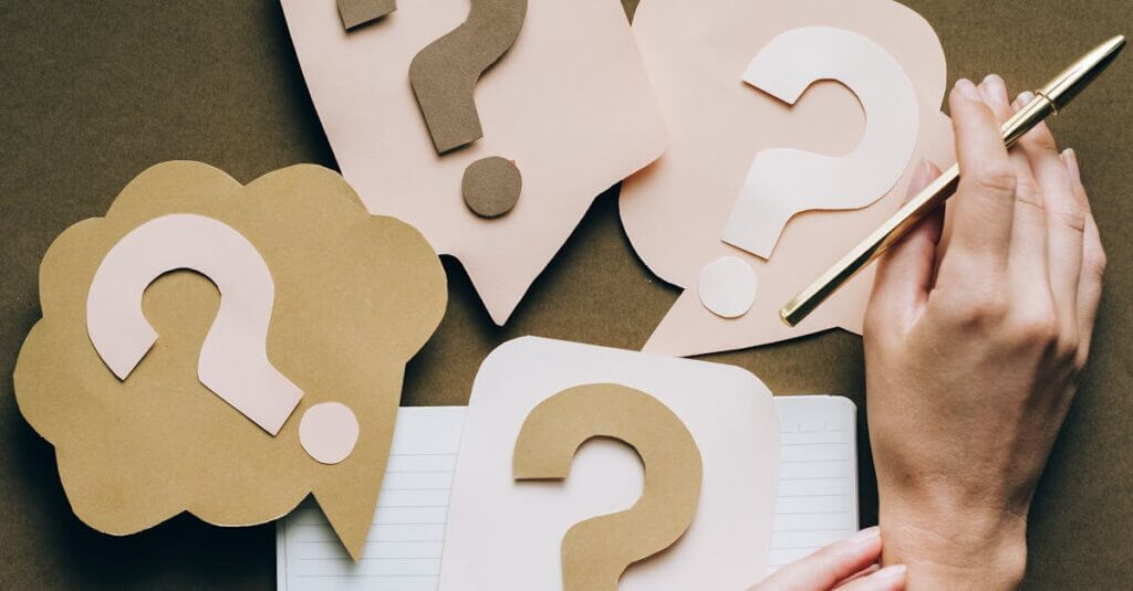 Hands holding pen next to paper question marks on notebook, symbolizing inquiry and creativity.
